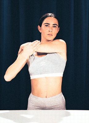 Esercizio per gli arti superiori. Movimenti per gli arti superiori da eseguire in piedi. Un braccio spinto fino a toccare con la mano la scapola opposta.